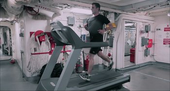 Movie still from “Aircraft Carrier: Guardian of the Seas” (2016), directed by Stephen Low – A man is running on a treadmill in a gym; Wide shot, High angle