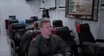 Movie still from “Aircraft Carrier: Guardian of the Seas” (2016), directed by Stephen Low – A man sitting in an airplane seat in front of a tv; Medium shot, High angle