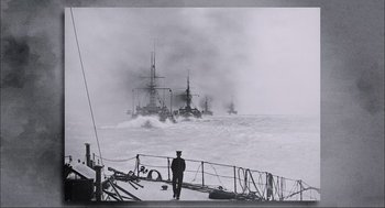 Movie still from “Aircraft Carrier: Guardian of the Seas” (2016), directed by Stephen Low – A black and white photo of a man standing on a boat in the water; Extreme Wide shot, High angle