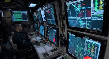 Movie still from “Aircraft Carrier: Guardian of the Seas” (2016), directed by Stephen Low – Many computer monitors on the ship; Medium shot, Over the shoulder angle