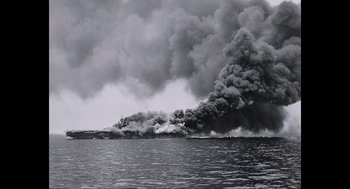 Movie still from “Aircraft Carrier: Guardian of the Seas” (2016), directed by Stephen Low – Smoke billows from a fire on the water; Extreme Wide shot, High angle