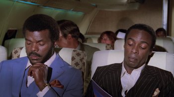 Movie still from “Airplane!” (1980), directed by David Zucker – Two men are sitting on a plane and one of them is holding a book; Close Up shot, Low angle
