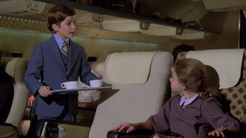 Movie still from “Airplane!” (1980), directed by David Zucker – A young boy in a business suit serving a woman on an airplane; Medium shot, High angle