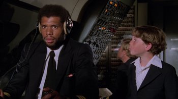 Movie still from “Airplane!” (1980), directed by David Zucker – A man in a suit and headphones in front of other people; Medium shot, Low angle