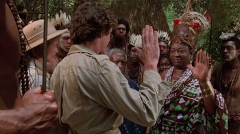Movie still from “Airplane!” (1980), directed by David Zucker – A group of people gathered around a man in native dress; Medium shot, Over the shoulder angle
