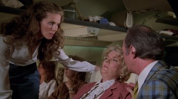 Movie still from “Airplane!” (1980), directed by David Zucker – A group of people sitting around a woman with her hand on her head; Medium shot, Over the shoulder angle