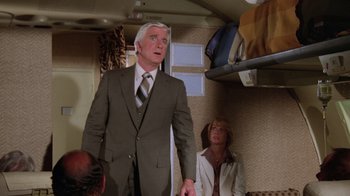 Movie still from “Airplane!” (1980), directed by David Zucker – An older man in a suit and tie standing next to a woman in a suit; Medium shot, Low angle