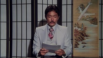 Movie still from “Airplane!” (1980), directed by David Zucker – A man sitting in front of a wall reading a paper; Medium shot, Over the shoulder angle