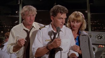 Movie still from “Airplane!” (1980), directed by David Zucker – A man in a pilot's uniform is holding a microphone while two other people look on; Medium shot, High angle
