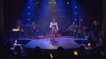 Movie still from “Alex Borstein: Corsets & Clown Suits” (2023), directed by Scott Ellis – A woman standing on a stage in front of a group of people; Extreme Wide shot, Over the shoulder angle