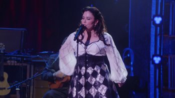 Movie still from “Alex Borstein: Corsets & Clown Suits” (2023), directed by Scott Ellis – A woman is singing into a microphone on a stage; Medium shot, Low angle