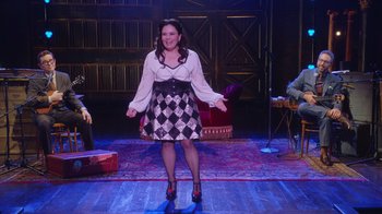 Movie still from “Alex Borstein: Corsets & Clown Suits” (2023), directed by Scott Ellis – A woman standing on a stage in front of a microphone; Wide shot, Over the shoulder angle