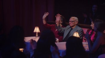Movie still from “Alex Borstein: Corsets & Clown Suits” (2023), directed by Scott Ellis – A man and a woman waving at a party; Wide shot, High angle