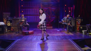 Movie still from “Alex Borstein: Corsets & Clown Suits” (2023), directed by Scott Ellis – A woman in a checkered skirt on a stage; Wide shot, Over the shoulder angle