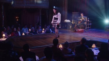 Movie still from “Alex Borstein: Corsets & Clown Suits” (2023), directed by Scott Ellis – A woman standing on a stage in front of a group of people; Wide shot, Over the shoulder angle