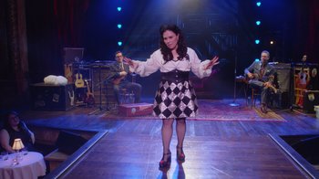 Movie still from “Alex Borstein: Corsets & Clown Suits” (2023), directed by Scott Ellis – A woman in a black and white dress on a stage; Wide shot, High angle