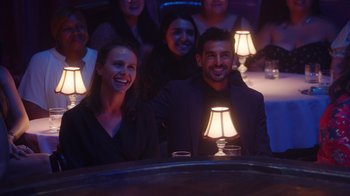 Movie still from “Alex Borstein: Corsets & Clown Suits” (2023), directed by Scott Ellis – A group of people sitting at a table in front of a lamp; Wide shot, Over the shoulder angle
