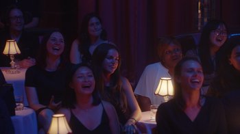 Movie still from “Alex Borstein: Corsets & Clown Suits” (2023), directed by Scott Ellis – A group of people sitting at a table with lamps; Medium shot, Over the shoulder angle