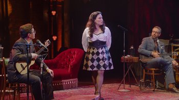 Movie still from “Alex Borstein: Corsets & Clown Suits” (2023), directed by Scott Ellis – A woman standing on a stage in front of a microphone; Wide shot, Over the shoulder angle