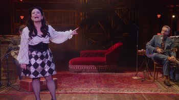 Movie still from “Alex Borstein: Corsets & Clown Suits” (2023), directed by Scott Ellis – A woman standing on a stage in front of a microphone; Wide shot, High angle