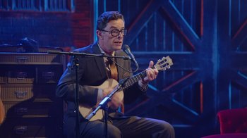 Movie still from “Alex Borstein: Corsets & Clown Suits” (2023), directed by Scott Ellis – A man sitting in front of a microphone holding a guitar; Medium shot, Over the shoulder angle
