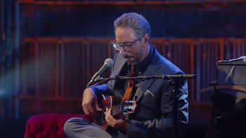 Movie still from “Alex Borstein: Corsets & Clown Suits” (2023), directed by Scott Ellis – A man in a suit and tie playing a ukulele; Medium shot, Low angle