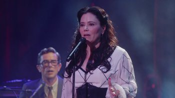 Movie still from “Alex Borstein: Corsets & Clown Suits” (2023), directed by Scott Ellis – A woman is singing into a microphone while a man looks on; Medium shot, Over the shoulder angle