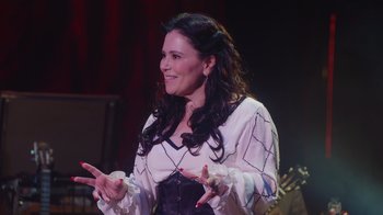 Movie still from “Alex Borstein: Corsets & Clown Suits” (2023), directed by Scott Ellis – A woman in a white shirt is smiling and clapping hands; Medium shot, Over the shoulder angle