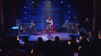 Movie still from “Alex Borstein: Corsets & Clown Suits” (2023), directed by Scott Ellis – A woman singing on a stage with a group of people watching; Wide shot, Over the shoulder angle