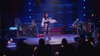 Movie still from “Alex Borstein: Corsets & Clown Suits” (2023), directed by Scott Ellis – A woman standing on a stage in front of a group of people; Wide shot, Over the shoulder angle
