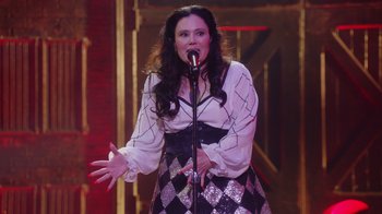 Movie still from “Alex Borstein: Corsets & Clown Suits” (2023), directed by Scott Ellis – A woman is singing into a microphone on a stage; Medium shot, Low angle