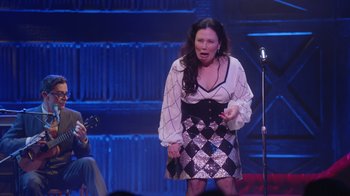 Movie still from “Alex Borstein: Corsets & Clown Suits” (2023), directed by Scott Ellis – A woman standing on a stage with a microphone; Medium shot, Over the shoulder angle