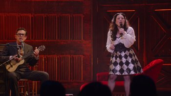 Movie still from “Alex Borstein: Corsets & Clown Suits” (2023), directed by Scott Ellis – A woman is singing into a microphone on a stage; Wide shot, High angle