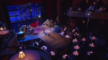 Movie still from “Alex Borstein: Corsets & Clown Suits” (2023), directed by Scott Ellis – An audience is sitting on a stage and a woman is singing; Extreme Wide shot, High angle