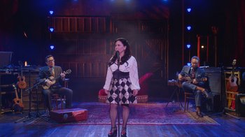 Movie still from “Alex Borstein: Corsets & Clown Suits” (2023), directed by Scott Ellis – A woman standing on a stage in front of two other people; Wide shot, High angle