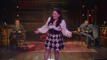 Movie still from “Alex Borstein: Corsets & Clown Suits” (2023), directed by Scott Ellis – A woman standing on a stage in front of a microphone; Wide shot, High angle