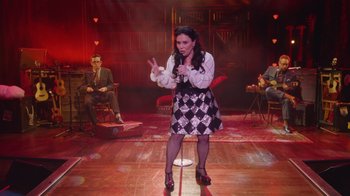 Movie still from “Alex Borstein: Corsets & Clown Suits” (2023), directed by Scott Ellis – A woman standing on a wooden floor in front of a microphone; Wide shot, High angle