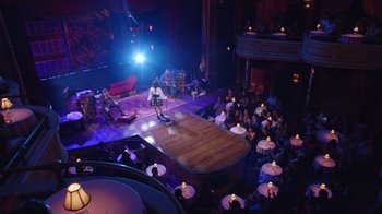 Movie still from “Alex Borstein: Corsets & Clown Suits” (2023), directed by Scott Ellis – An audience is sitting on a stage and a man is standing on the stage; Extreme Wide shot, High angle