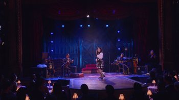 Movie still from “Alex Borstein: Corsets & Clown Suits” (2023), directed by Scott Ellis – A group of people sitting on a stage playing instruments and singing; Extreme Wide shot, Over the shoulder angle