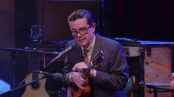Movie still from “Alex Borstein: Corsets & Clown Suits” (2023), directed by Scott Ellis – A man in a suit and tie playing a ukulele; Medium shot, Over the shoulder angle