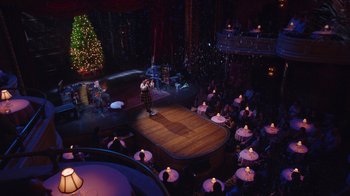 Movie still from “Alex Borstein: Corsets & Clown Suits” (2023), directed by Scott Ellis – An audience is sitting in a circle in front of a christmas tree; Extreme Wide shot, High angle