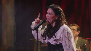 Movie still from “Alex Borstein: Corsets & Clown Suits” (2023), directed by Scott Ellis – A woman with long brown curly hair holding a microphone; Medium shot, Over the shoulder angle