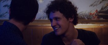 Movie still from “Alex Strangelove” (2018), directed by Craig Johnson – A man with curly black hair smiling at a party; Close Up shot, Over the shoulder angle