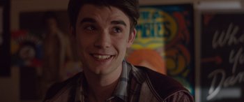 Movie still from “Alex Strangelove” (2018), directed by Craig Johnson – A young man smiling for the camera in front of posters; Close Up shot, Over the shoulder angle