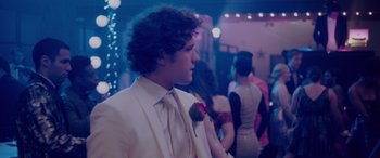 Movie still from “Alex Strangelove” (2018), directed by Craig Johnson – A man in a white suit with a red flower in his lapel; Medium shot, Over the shoulder angle