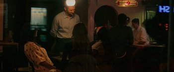 Movie still from “Alice, Darling” (2022), directed by Mary Nighy – A group of people sitting in a room with a man wearing a hat; Medium shot, Over the shoulder angle