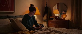 Movie still from “Alice, Darling” (2022), directed by Mary Nighy – A woman sitting on top of a bed looking at a tablet; Medium shot, High angle