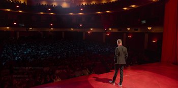 Movie still from “All About Gila” (2022), directed by Arturo González-Campos – A man standing on a stage in front of an audience; Extreme Wide shot, High angle