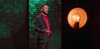 Movie still from “All About Gila” (2022), directed by Arturo González-Campos – A man in a red shirt and black suit standing in front of a green background; Medium shot, Low angle