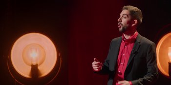 Movie still from “All About Gila” (2022), directed by Arturo González-Campos – A man standing in front of a microphone on a stage; Medium shot, Over the shoulder angle
